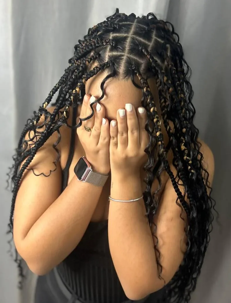 Boho knotless braids with caramel highlights and curly ends; the model covers her face with white-manicured hands in front of a grey curtain