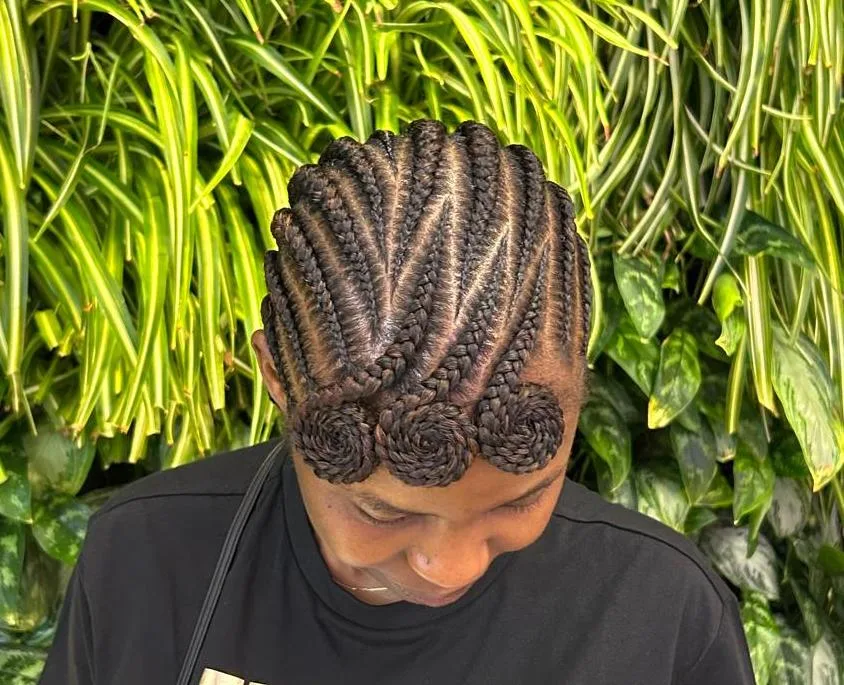 Overhead view of stitch-braid cornrows finished with three spiral buns, against a green plant wall