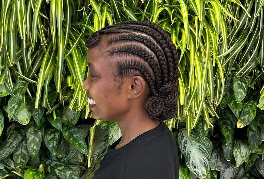 Smiling side profile showing cornrows gathered into a low spiral bun, in front of a plant wall
